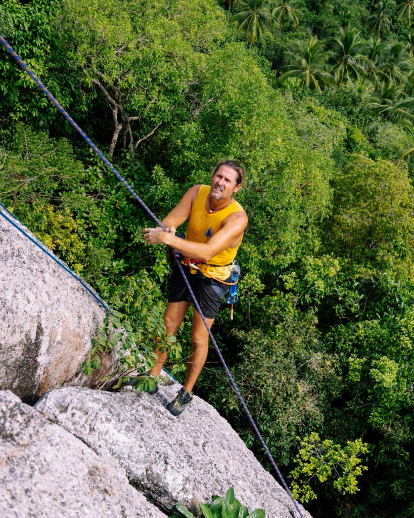 A man climbing a rocky mountain amidst lush green jungle using ropes for safety outdoors.