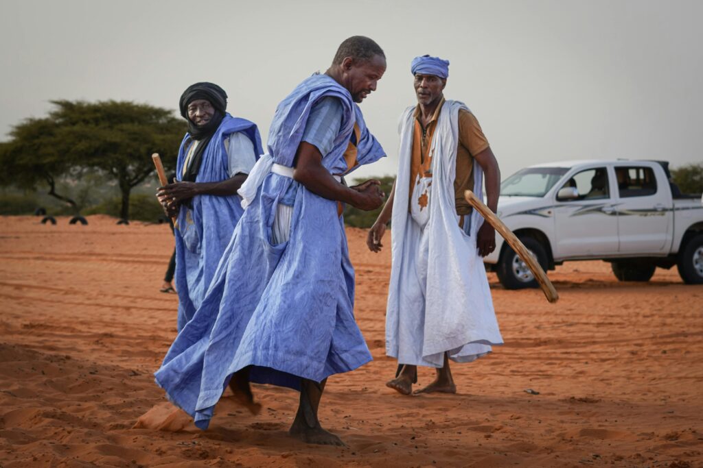 Three men in traditional blue robes engage in a cultural dance in a desert landscape.