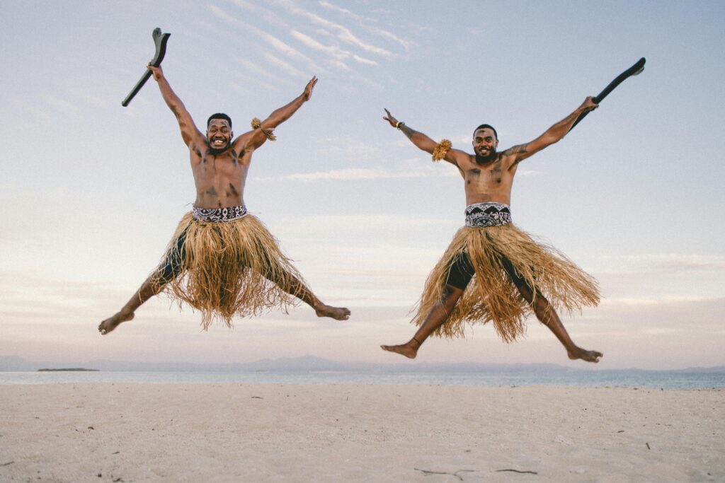 Joyful Fijian men in traditional skirts leaping on a beach with the ocean in the background.