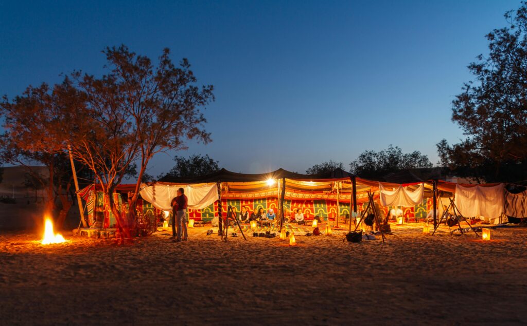 A vibrant desert camp in Taghit, Algeria, illuminated at dusk with a campfire and traditional decorations.