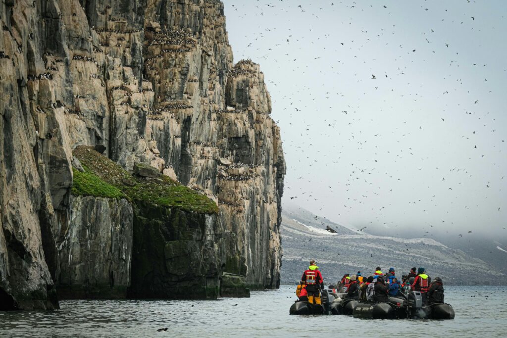 Visitors in boats observe seabirds on steep cliffs in an Arctic landscape.