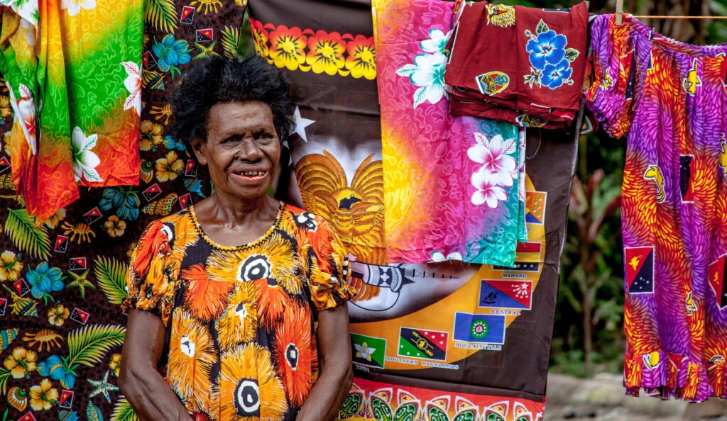 A senior woman stands smiling amidst vibrant traditional Papua New Guinean clothing.