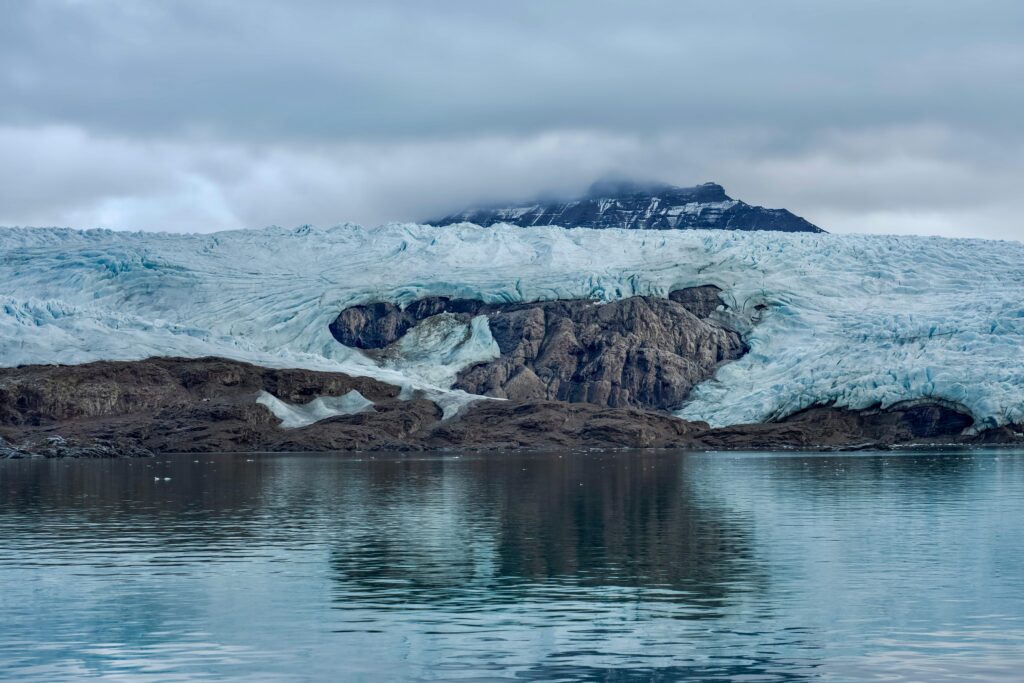 A stunning view of an arctic glacier with rugged cliffs and reflective icy waters under a cloudy sky.