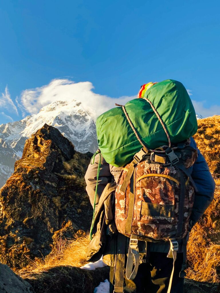 Back view of unrecognizable explorer with backpacks in warm clothes standing on stony ground and admiring amazing scenery of mountain ridge under cloudless blue sky