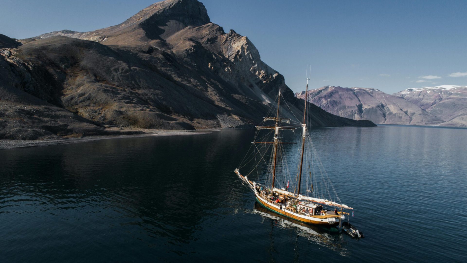 A schooner sails along a dramatic coastline with towering rock mountains under a clear sky.
